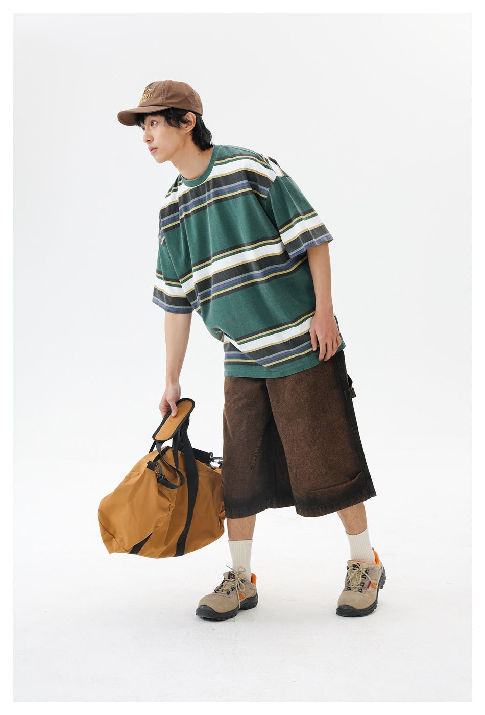 Model leans forward in a forest-green oversized T-shirt with broad white and narrow multicolor stripes, holding a tan duffel