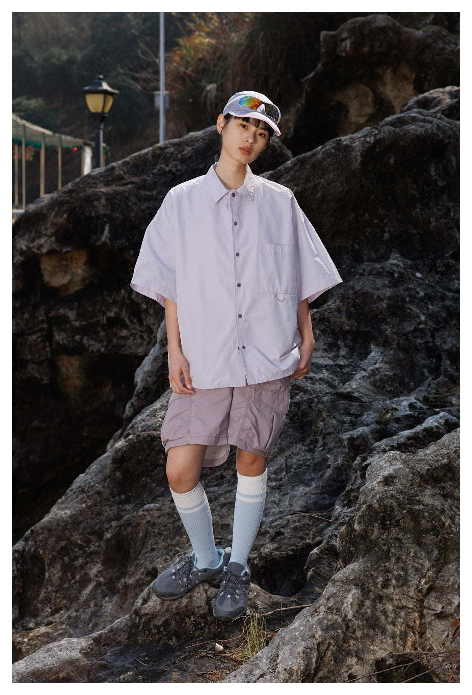 Model wearing an oversized pale lavender short-sleeve button-up shirt with mauve shorts, knee socks, and a cap.