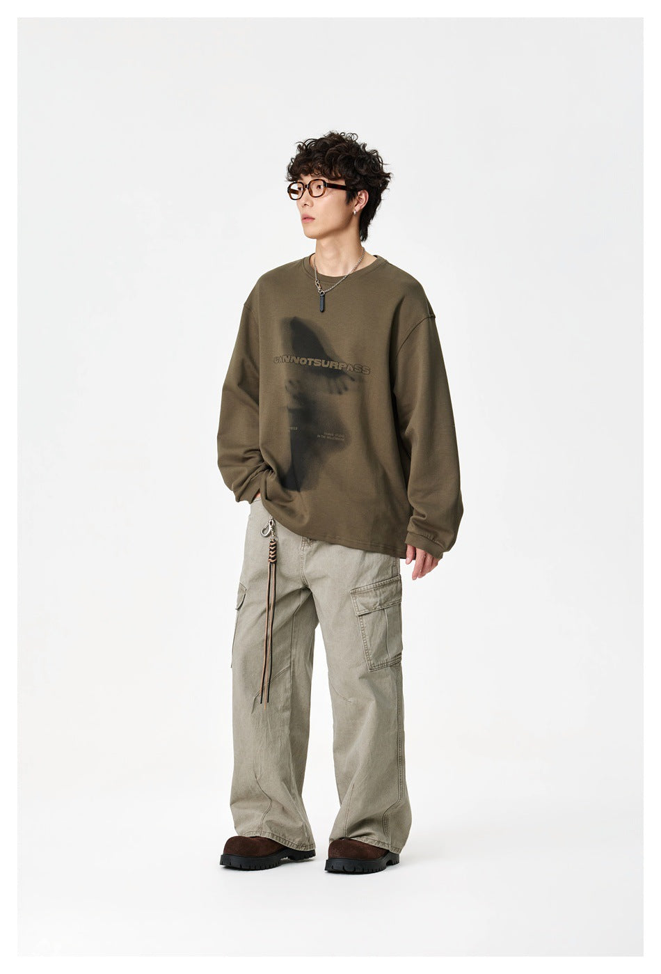 Model wearing an olive oversized sweatshirt with a dark graphic and text print, paired with light cargo pants.