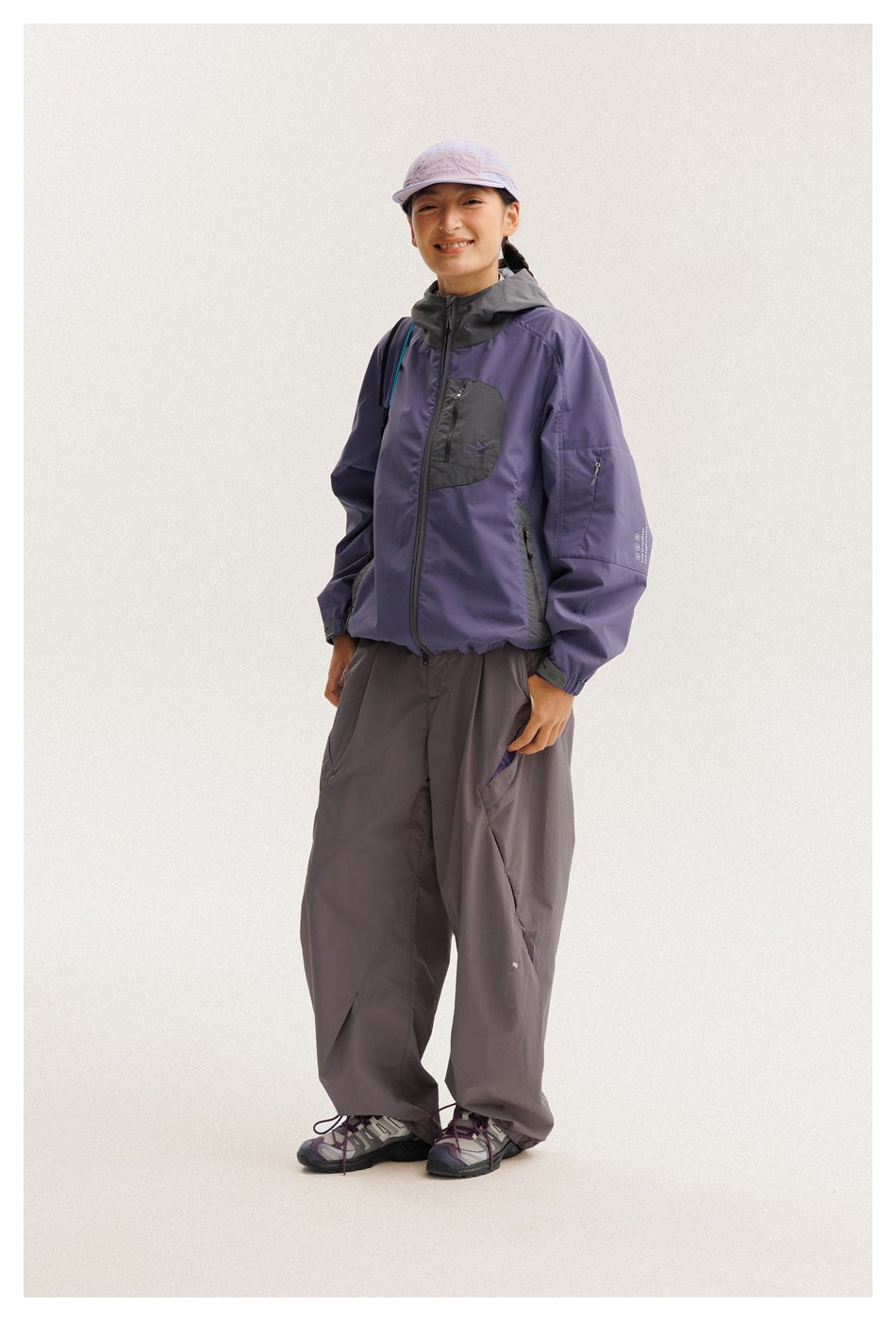 Model wearing a purple hooded jacket with charcoal panels, loose gray pants, and a lavender cap, shown full length.
