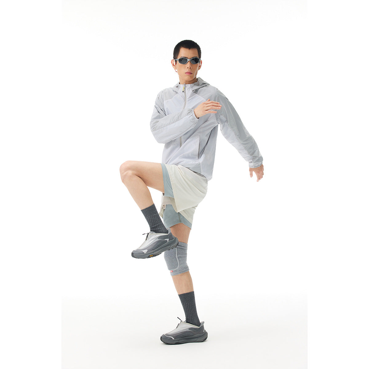 Man wearing a light gray hooded jacket with layered athletic shorts, sunglasses and gray sports accessories.