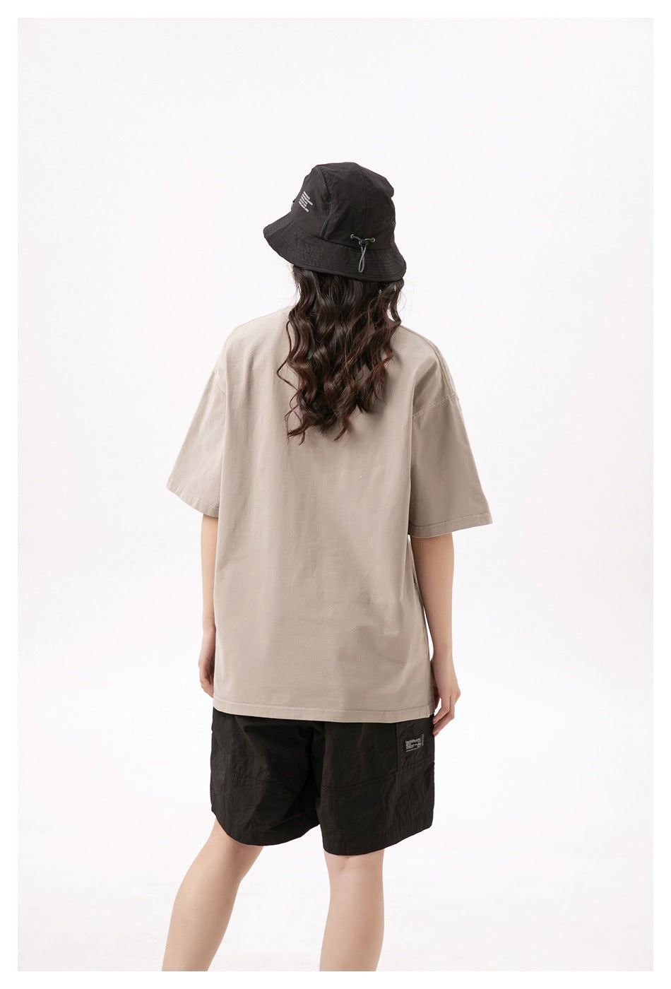 Back view of a model wearing a loose beige T-shirt, black shorts, and a black bucket hat.