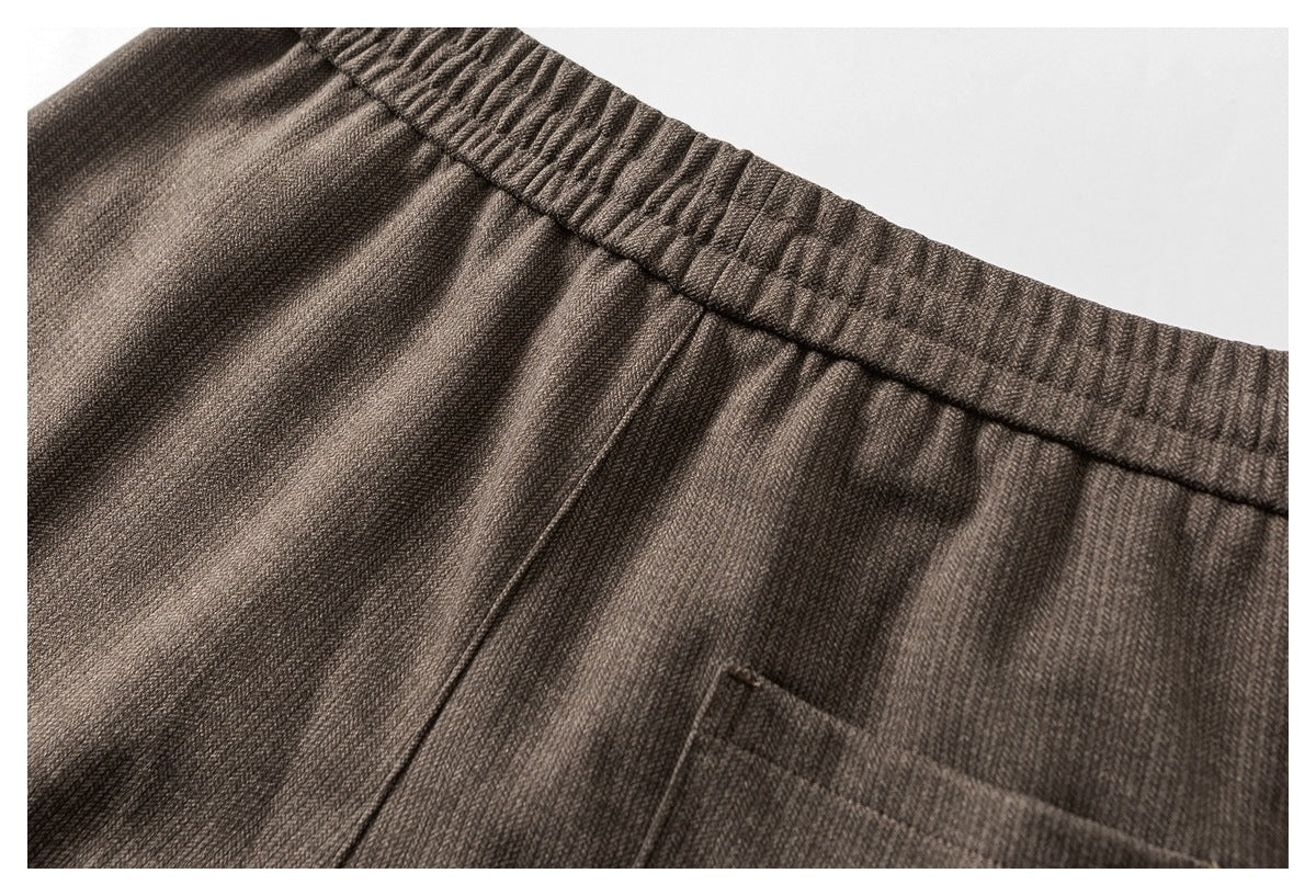 Close-up of taupe trousers showing the elastic waistband, front pleats, and pocket edge.