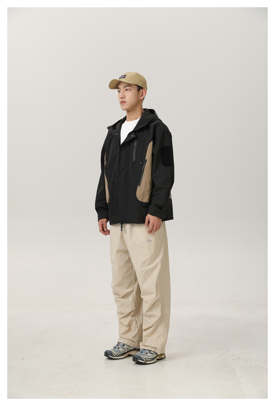 Man wearing a black and khaki hooded jacket over a white tee with beige pants, standing in a three-quarter view