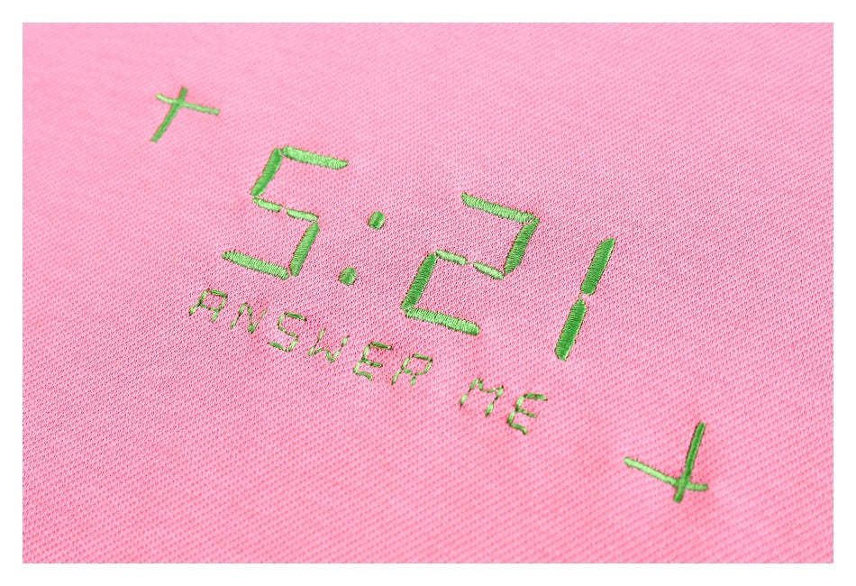 Close-up of green “5:21 ANSWER ME” embroidery and crosses on pink sweatshirt fabric.