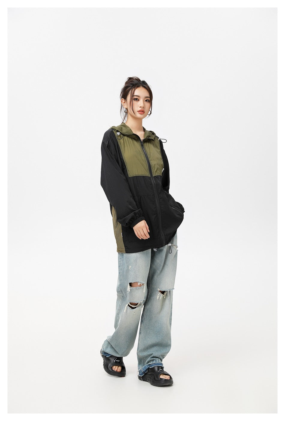Three-quarter view of a model wearing a black and olive hooded windbreaker with loose distressed jeans.