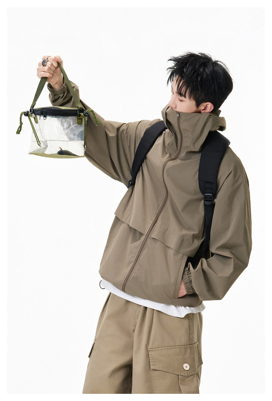 Model wearing an olive-brown hooded zip jacket and tan cargo shorts, holding a clear bag.