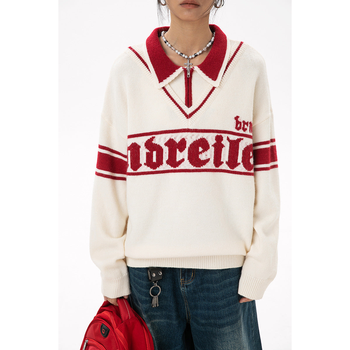 Close view of an ivory sweater on a model, showing its red zip collar, striped trim and large chest lettering.
