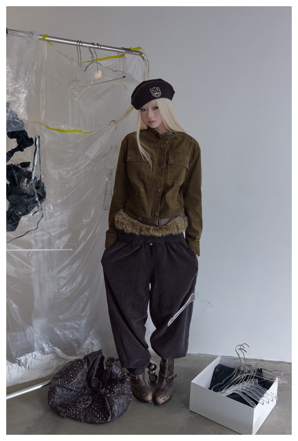 Model wearing the olive corduroy cropped jacket with beret, dark loose pants and boots in a studio with clear plastic