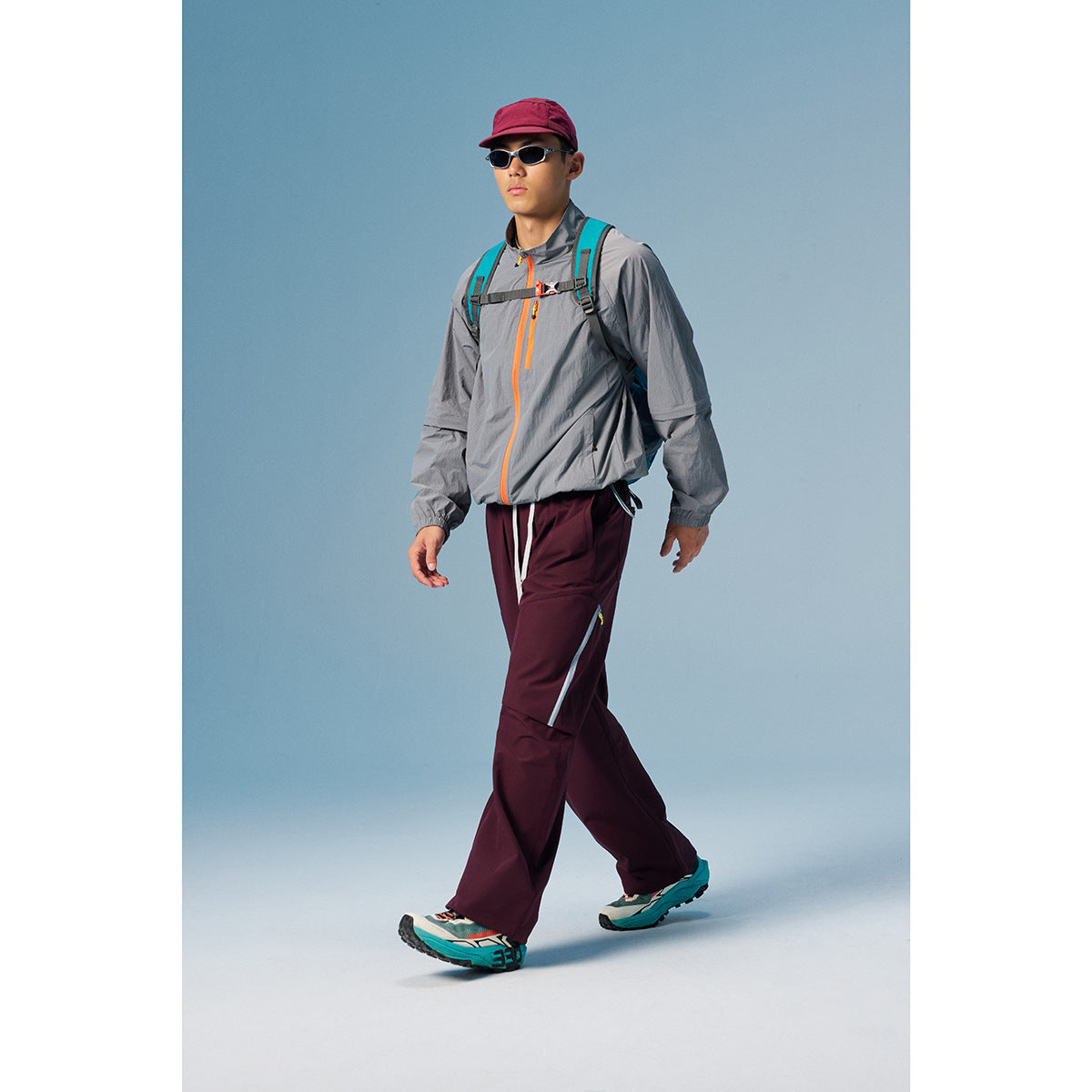 Man walking in a gray jacket with orange zippers and burgundy pants, wearing a backpack and cap.