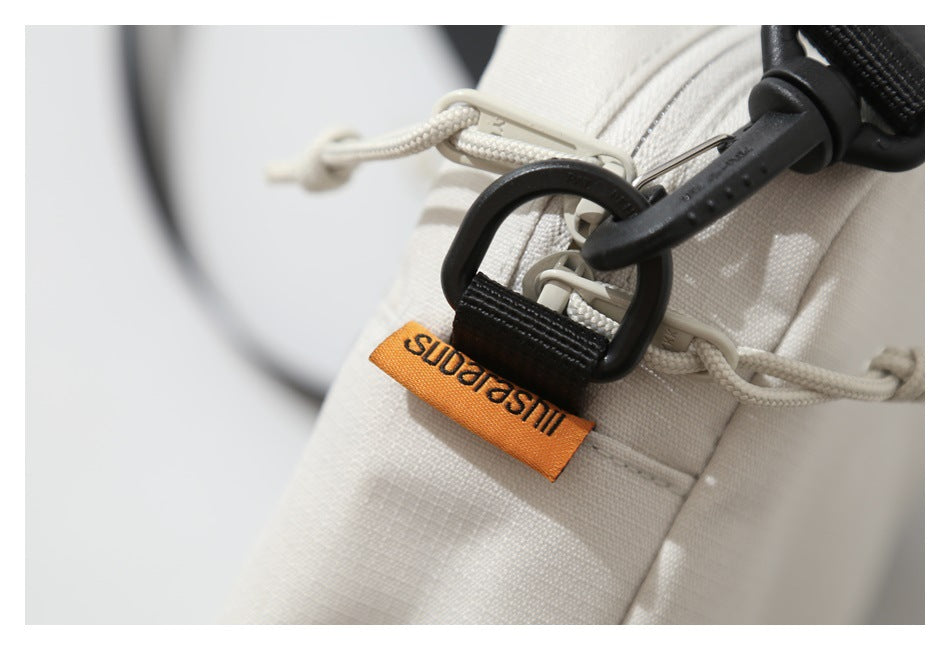 Close-up of a cream bag's drawcord, zipper pulls, black strap clip, and orange fabric label.