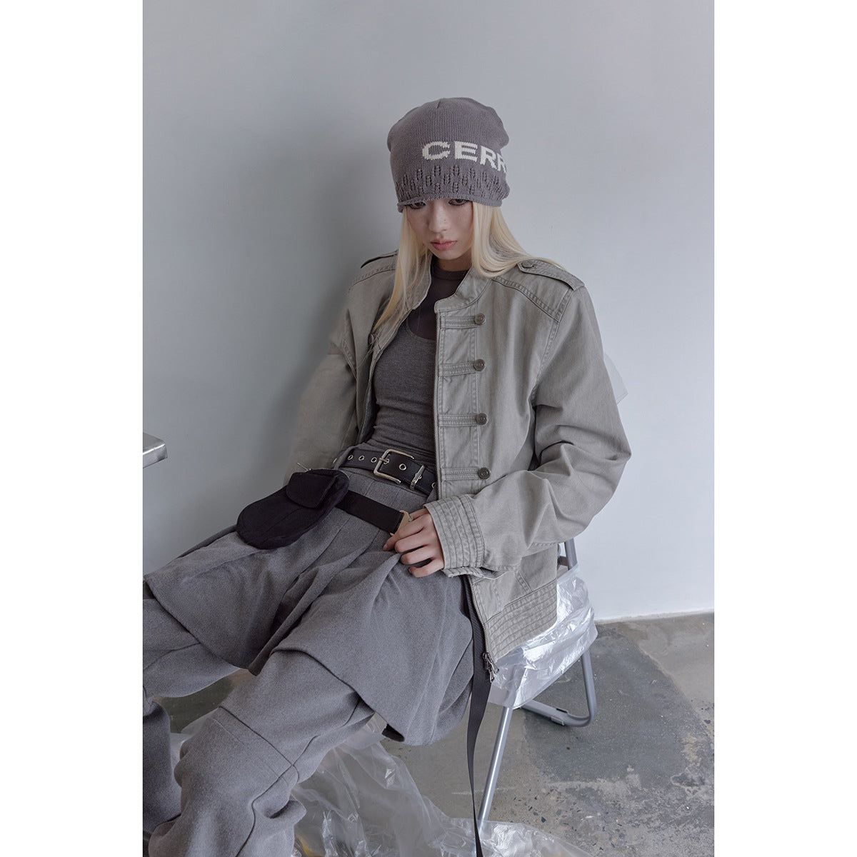 Seated model wearing a light gray military-style jacket with button tabs, a front zipper, and shoulder epaulettes.
