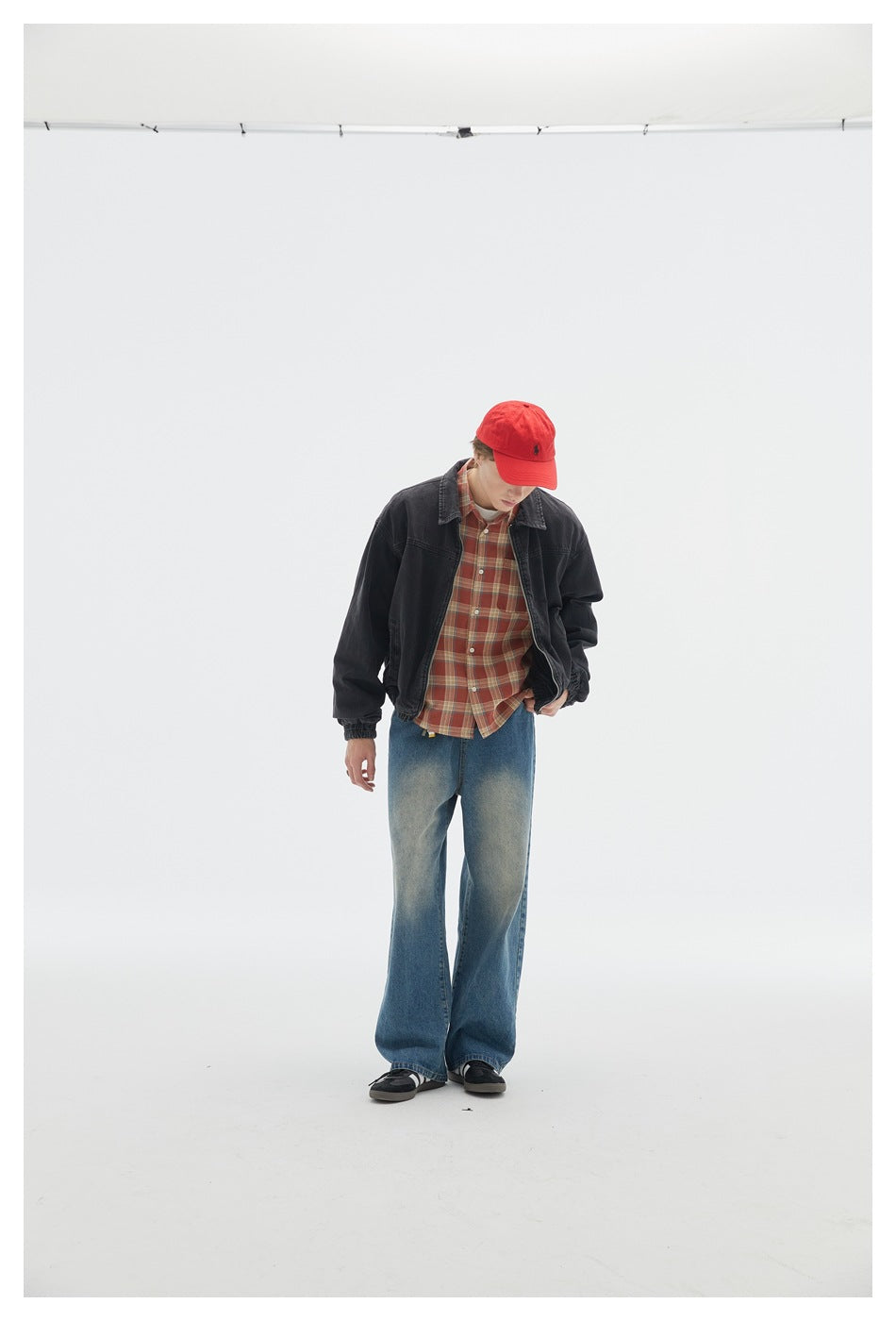 Person wearing a dark gray denim zip jacket over a plaid shirt with red cap and wide-leg jeans, looking down