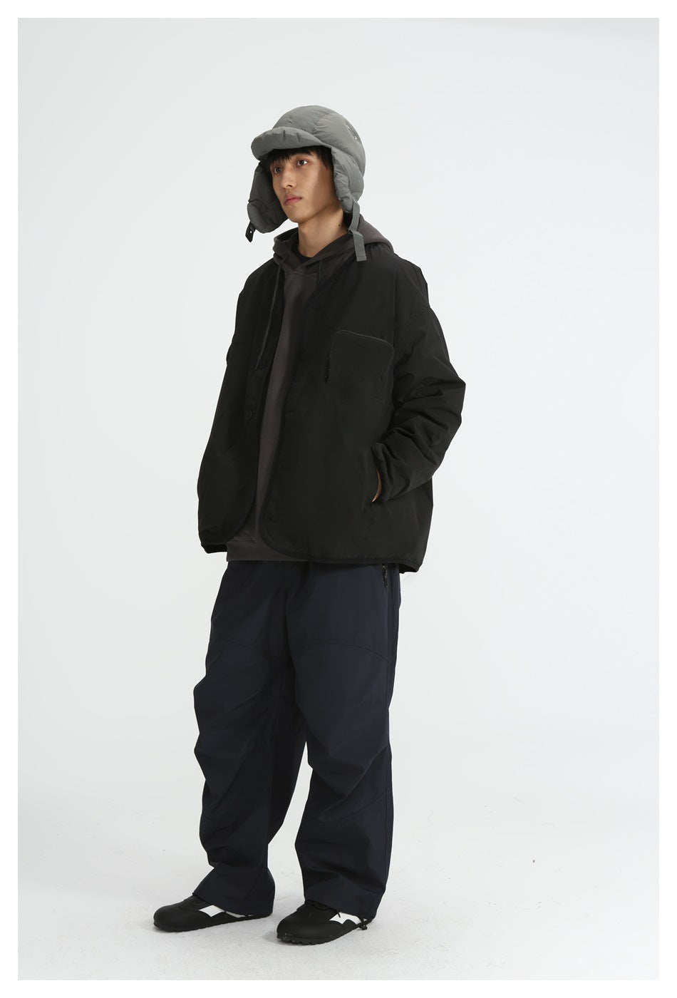 Model wearing a black collarless jacket over a dark hoodie with navy wide pants and a gray padded cap