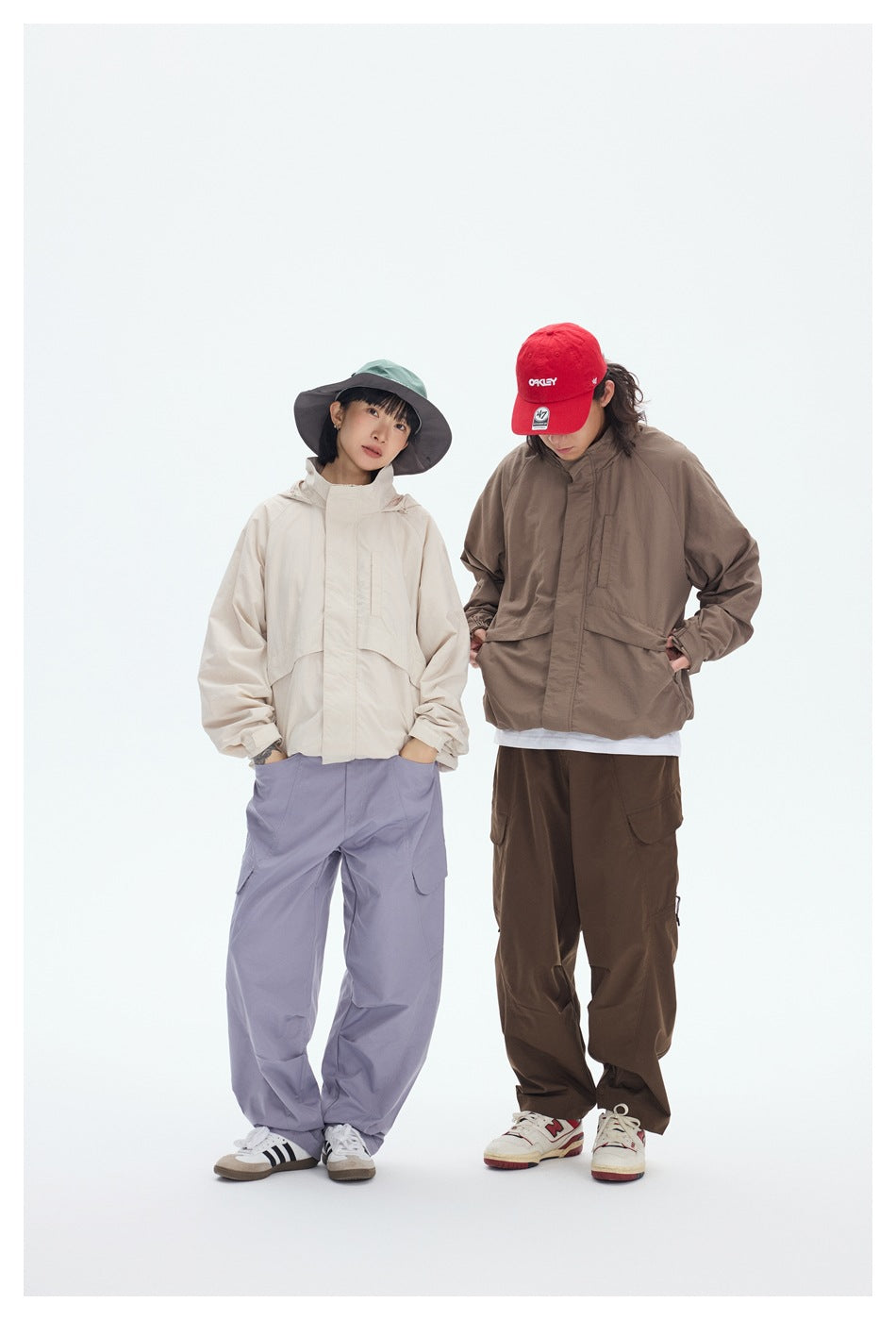 Two models wear a cream and a taupe hooded jacket with loose cargo pants; one wears a red cap.