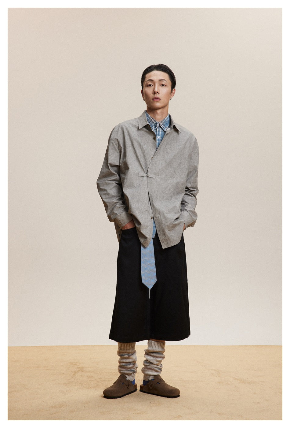 Model wearing a grey crinkled long-sleeve shirt over a plaid shirt with a black skirt and clogs