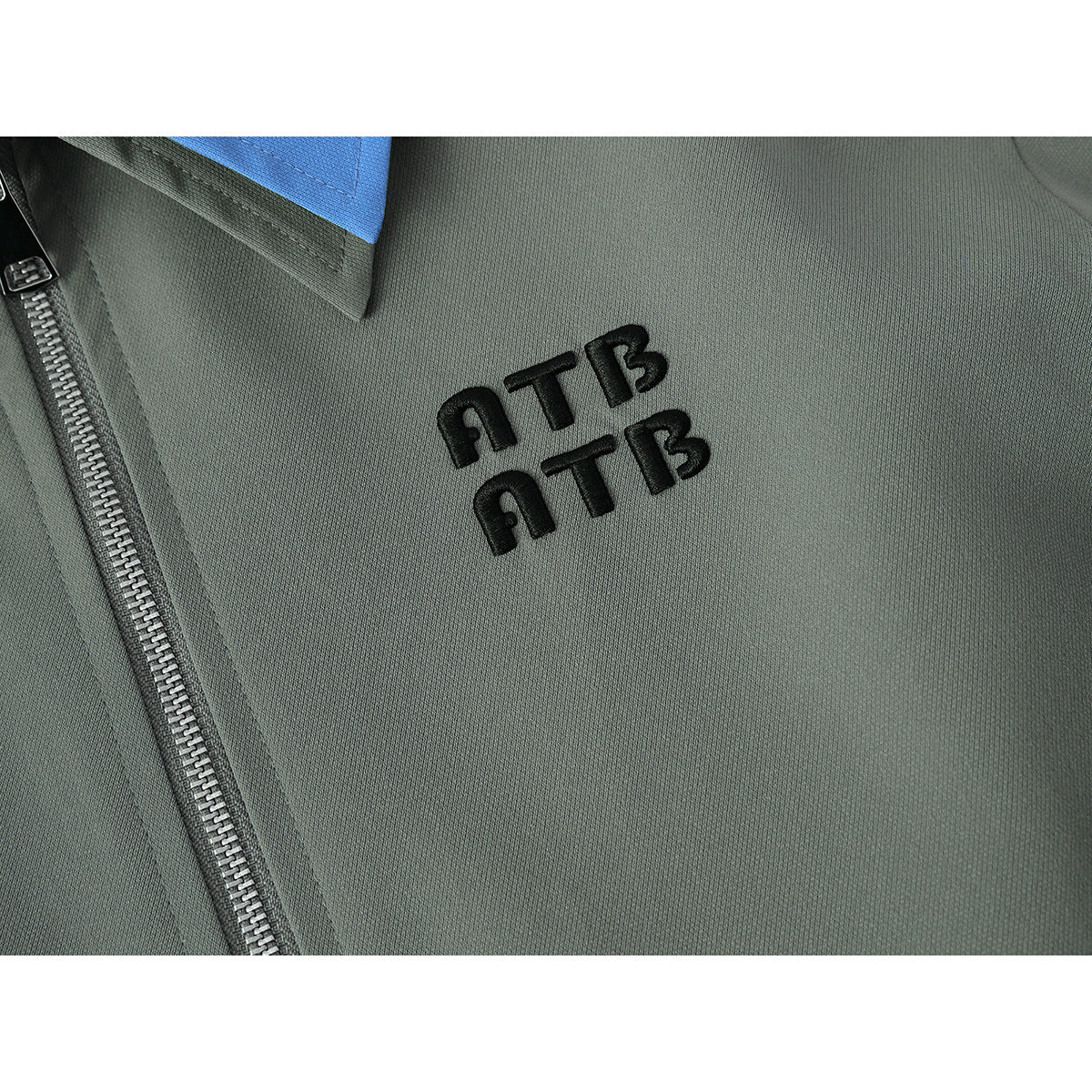 Close-up of black embroidered lettering on the gray-green jacket beside its silver front zipper.