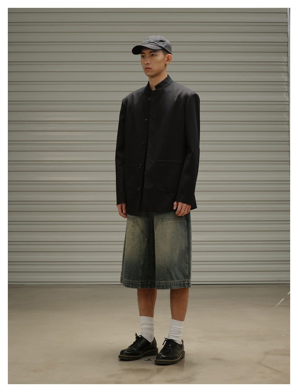 Three-quarter view of a model wearing a black band-collar coat, oversized faded denim shorts and a gray cap.