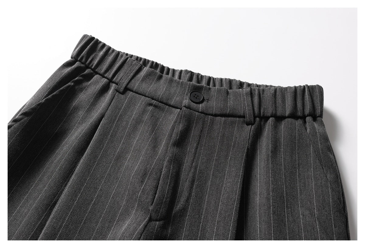 Close-up of charcoal pinstripe trousers showing the gathered waistband, belt loops, button, and front pleats.