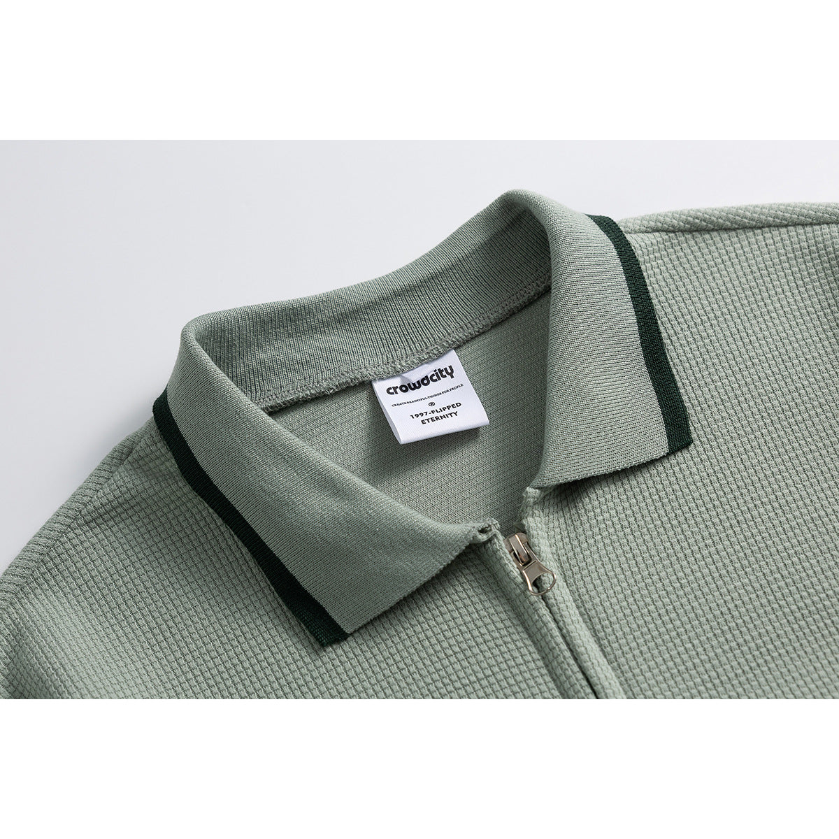 Close-up of a sage-green textured zip shirt showing its pointed collar, dark trim, zipper, and neck label.