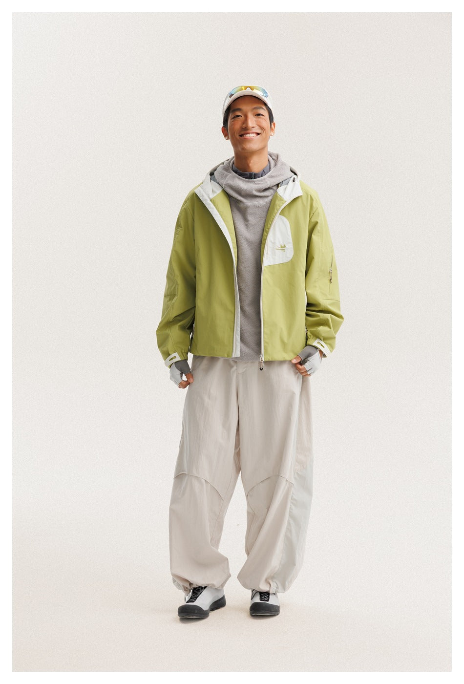 Model wears a lime green jacket with white trim and chest pocket, a gray top, and loose off-white pants.