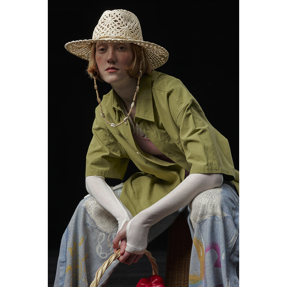 Seated model wearing an oversized olive-green short-sleeve collared shirt over a pink top.