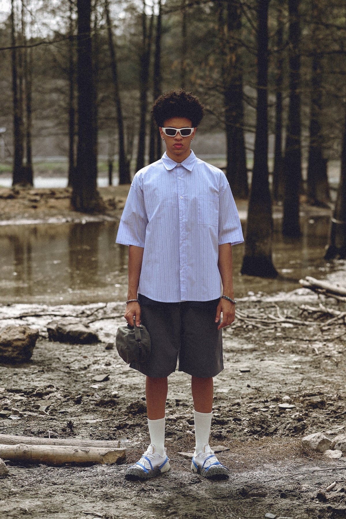 Person wearing a light blue striped short-sleeve shirt with gray shorts, white socks and sneakers in a forest by water