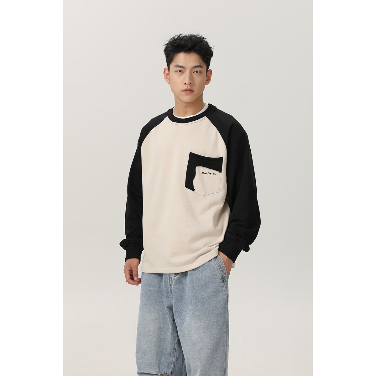 Front view of a model wearing a cream sweatshirt with black raglan sleeves, a contrasting chest pocket, and light-wash jeans.