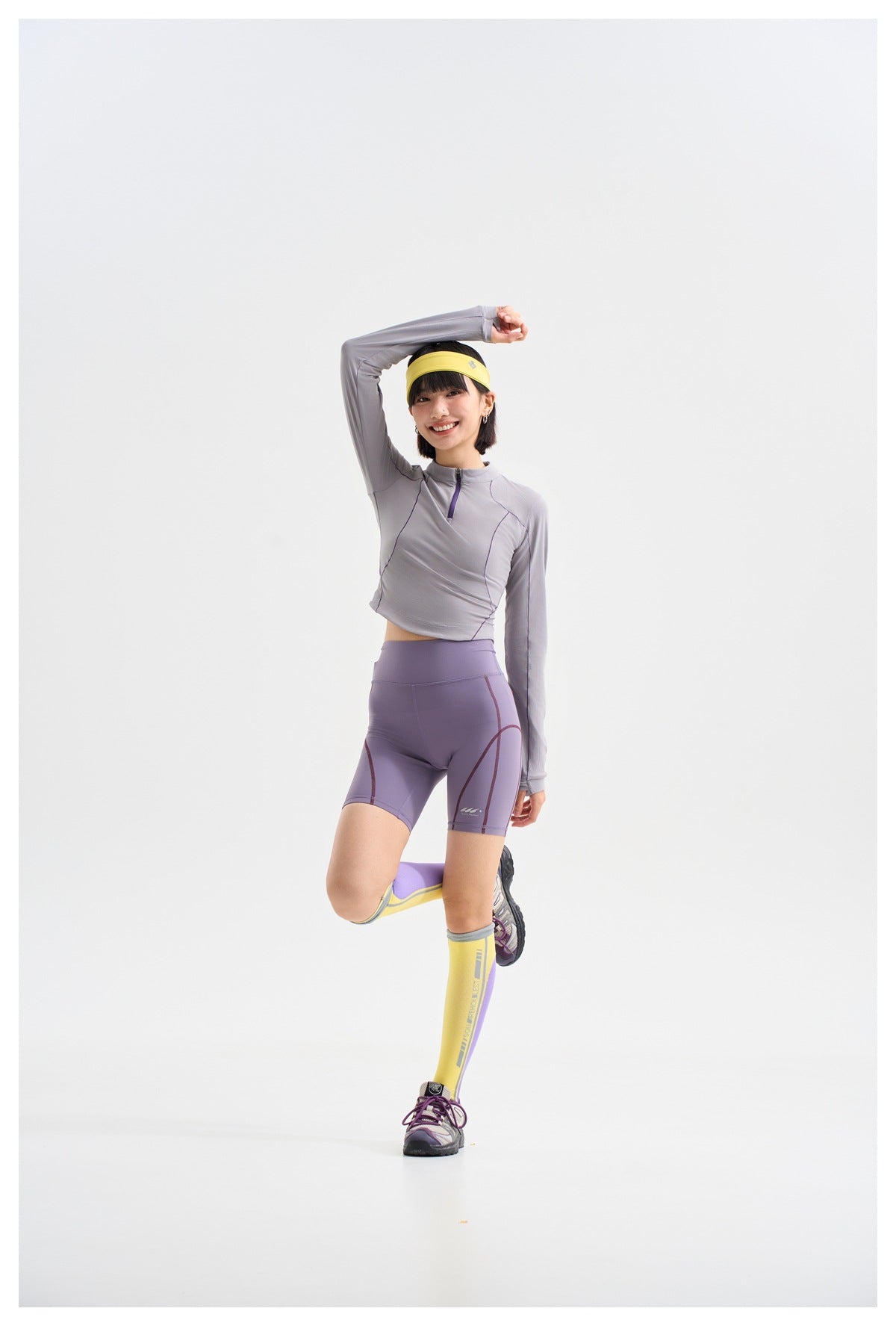 Model posing in a light gray-lavender quarter-zip top, lavender cycling shorts, and yellow-and-purple knee-high socks.