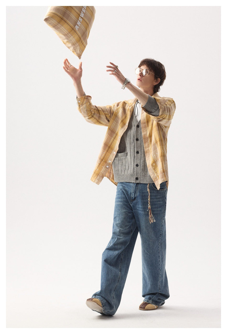 Model in a tan plaid shirt and gray cardigan reaches toward a matching plaid cushion in the air.