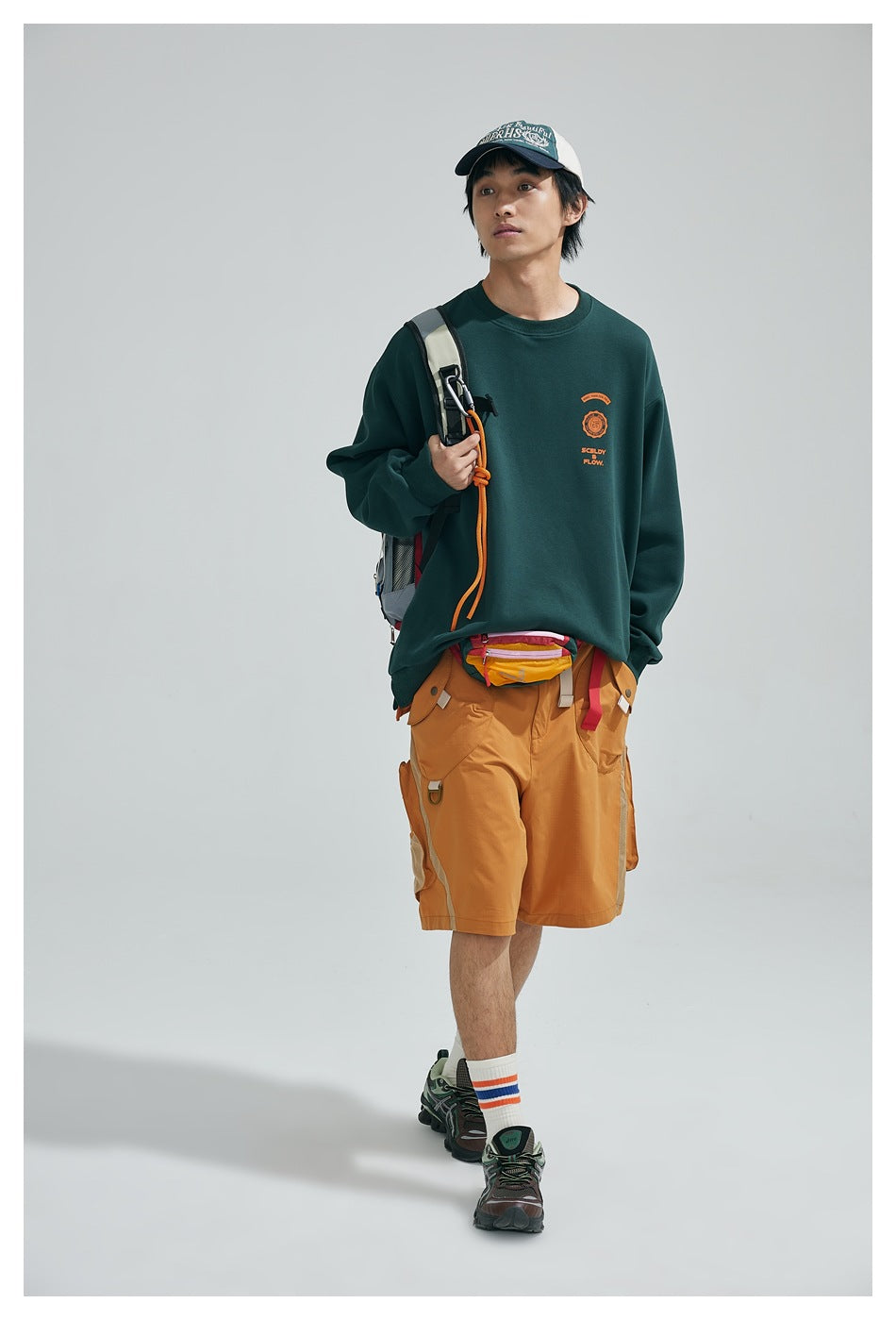 Model wearing a dark green crewneck sweatshirt with a small orange chest graphic and mustard cargo shorts, carrying a