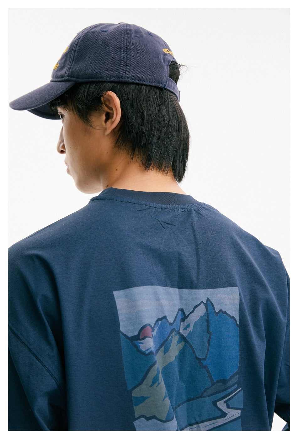 Close-up of a navy shirt's back mountain graphic, with part of the model and purple cap visible.