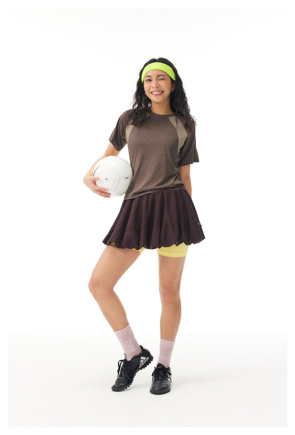 Full-length model wearing a brown paneled athletic top, dark flared skirt over yellow shorts, neon headband and holding a