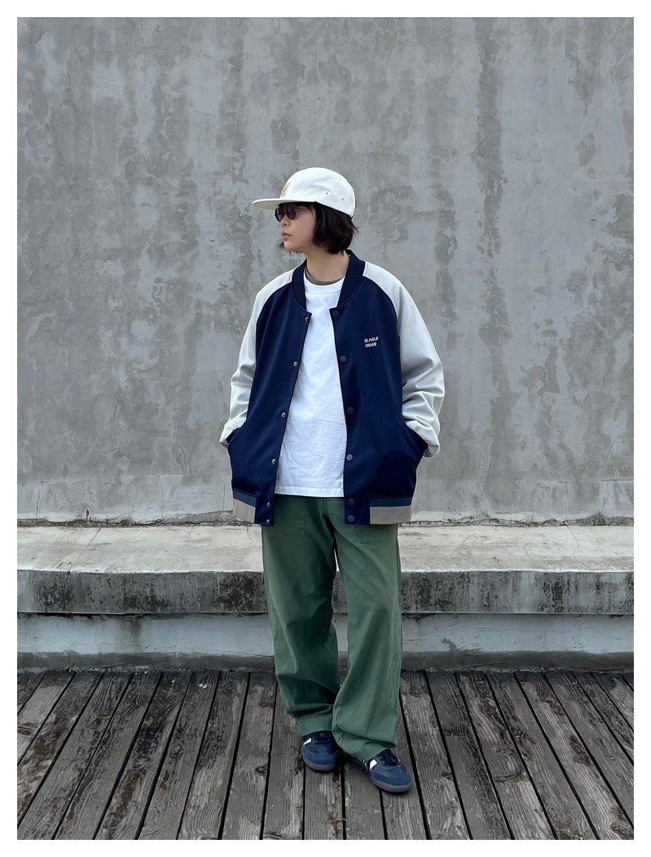 Person wearing navy and gray-green varsity jacket over a white tee with green pants against a concrete wall