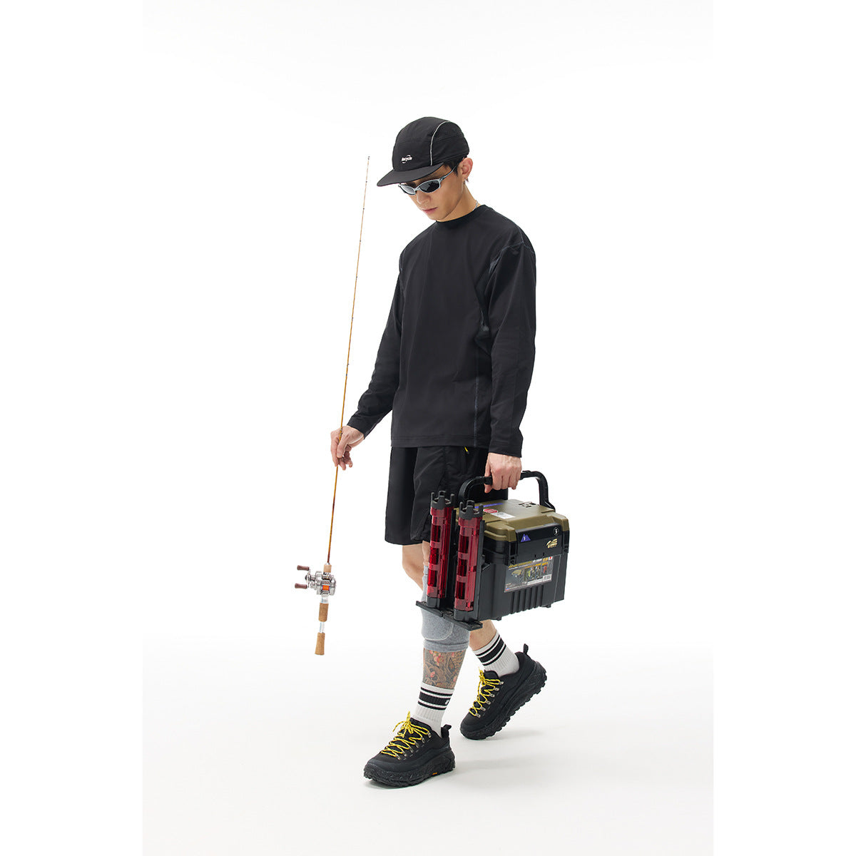 Model wearing a black long-sleeve shirt and shorts, holding a fishing rod and tackle box; front three-quarter view.