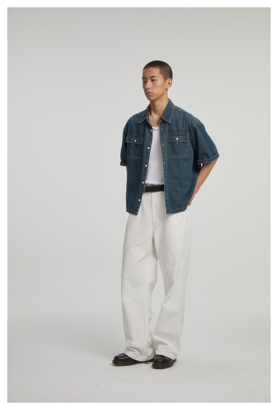 Full-length view of a model wearing a dark blue short-sleeve denim shirt with two chest pockets and white trousers.