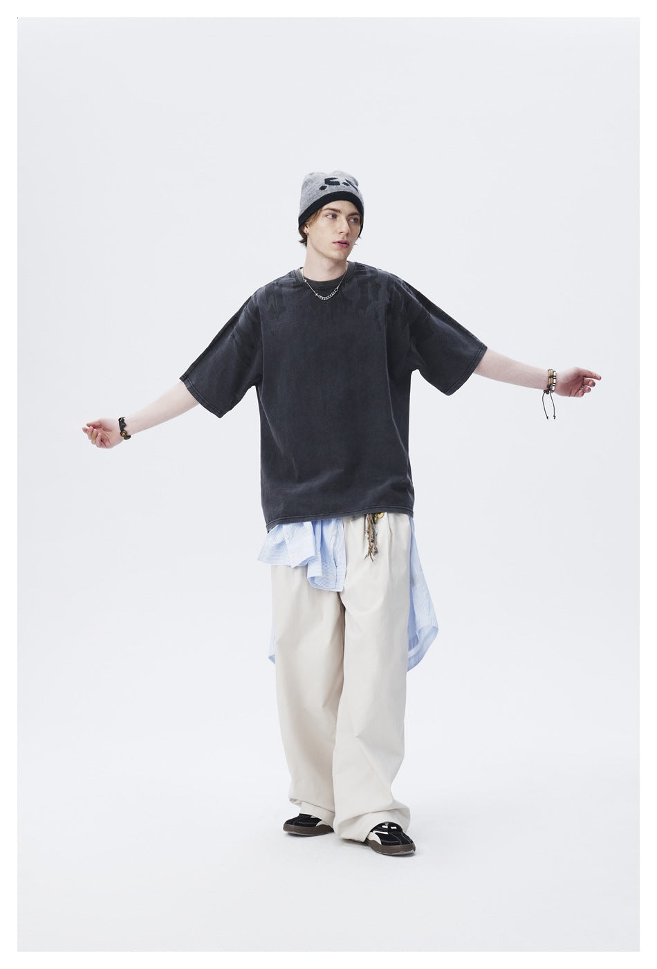 Full-length model in an oversized charcoal T-shirt with tonal shoulder graphics and loose cream trousers, arms outstretched.
