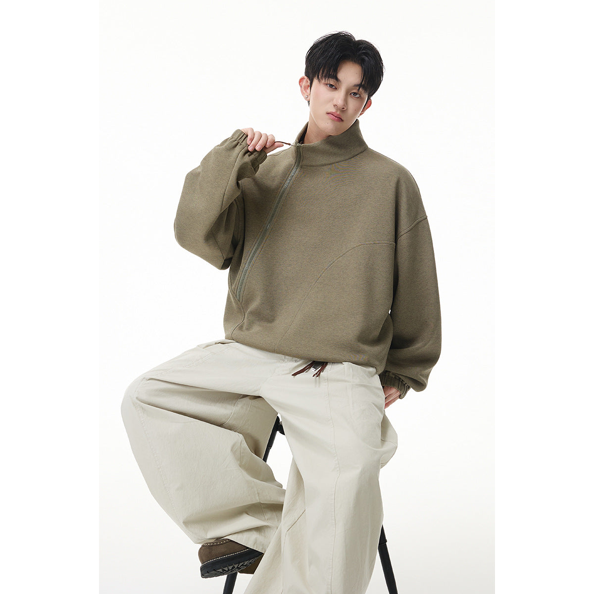 Model wearing an olive-gray oversized pullover with an asymmetrical zipper, tall collar, and cream trousers.