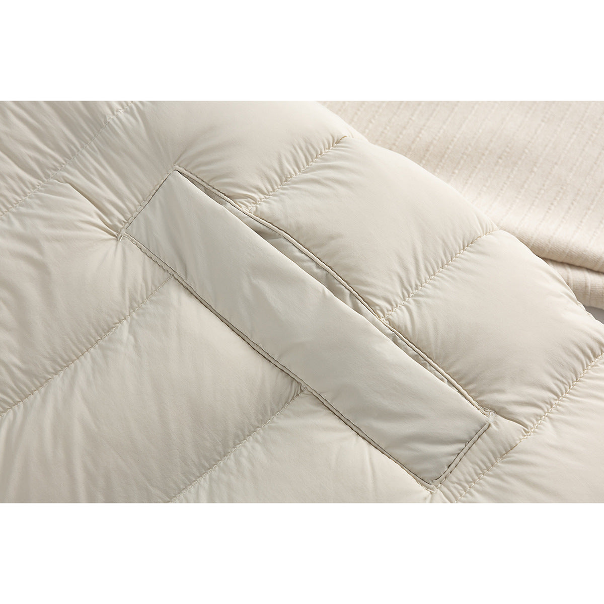Close-up of a cream quilted jacket showing a diagonal welt pocket opening and stitched panels.