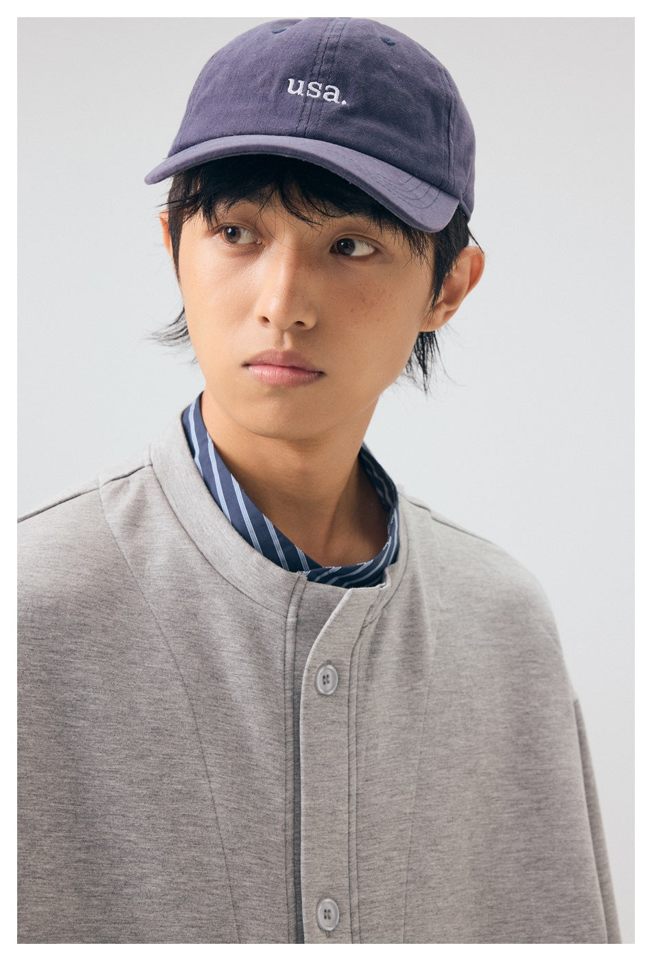 Close-up of a model wearing a light gray button-front jacket, striped shirt, and purple cap.