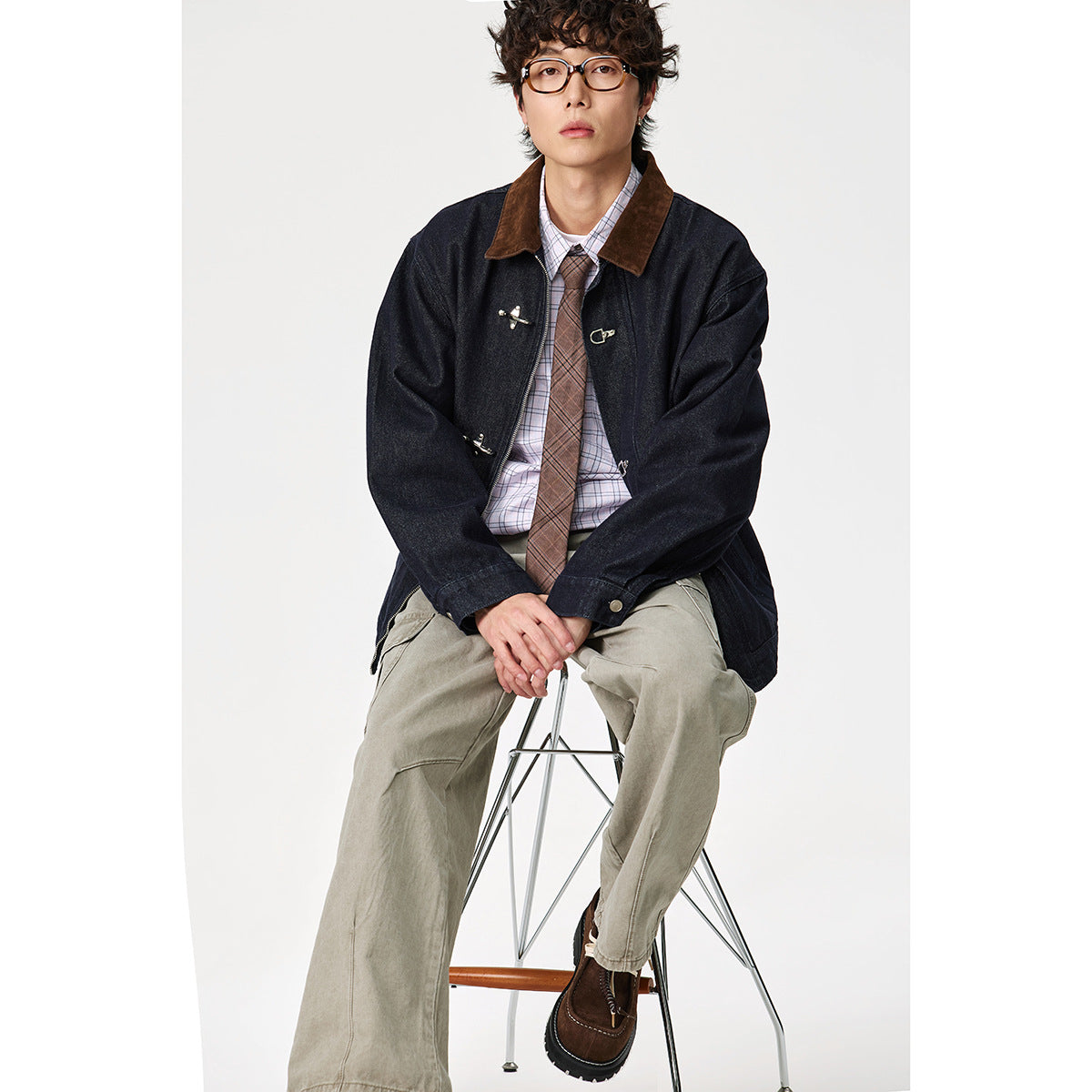 Seated model wearing a dark indigo jacket with a brown collar and silver toggle closures, styled with a plaid shirt and khaki