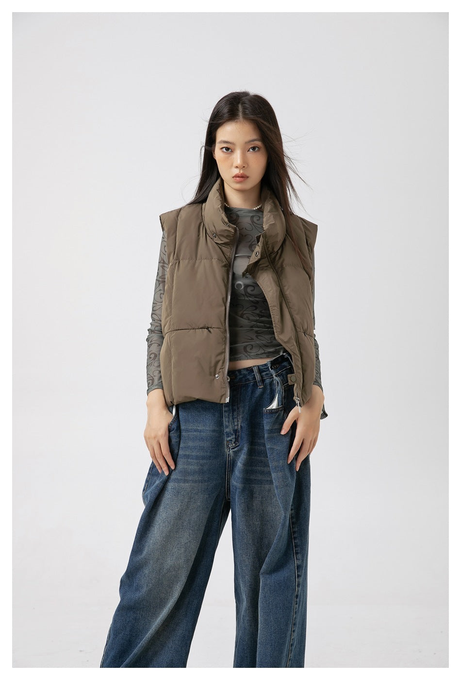 Woman wearing a cropped gray-green quilted stand-collar vest, zipped, with a patterned top and blue wide-leg jeans