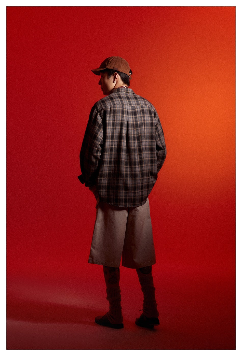 Model viewed from behind wearing a dark brown plaid button-up shirt, khaki shorts, brown cap, socks and shoes.