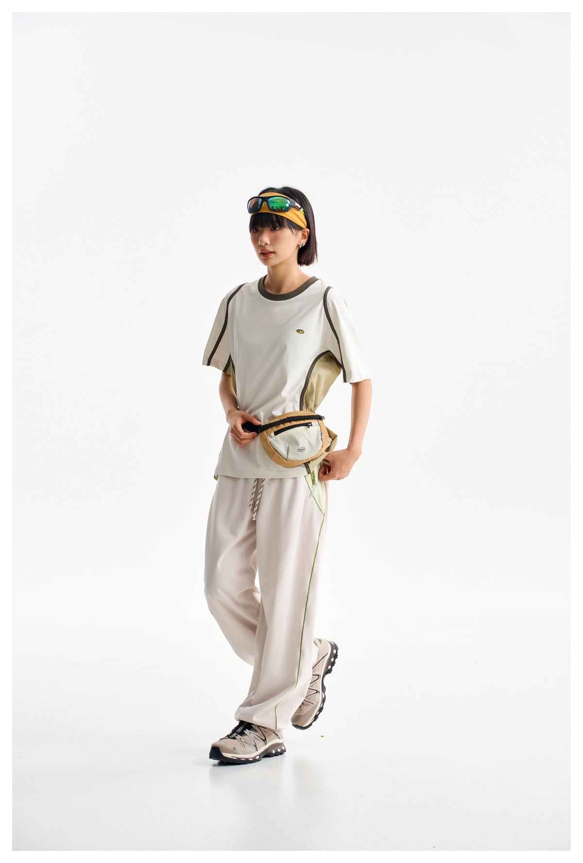 Model wearing an off-white piped T-shirt, loose white pants, a tan waist bag, and trail shoes.