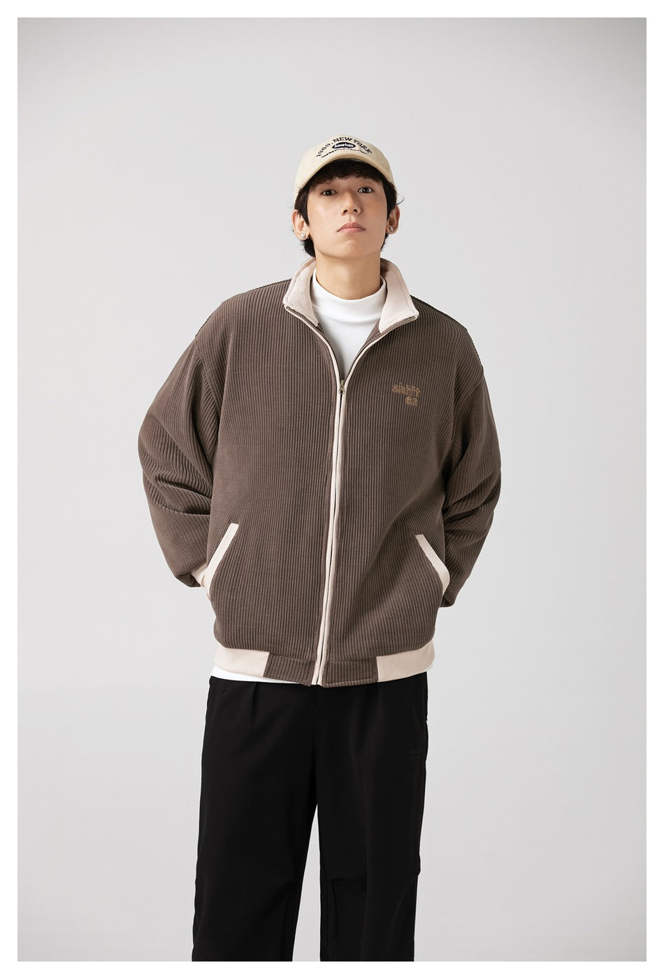 Model wearing brown ribbed zip jacket with cream trim over a white mock-neck top, black pants and a cream cap