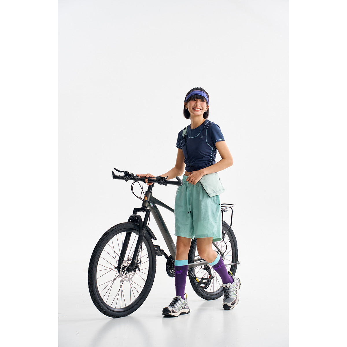 Model standing with a bicycle in a navy contrast-stitched tee, mint shorts and purple knee-high socks.