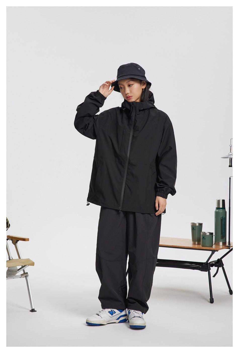 Full-length front view of a woman wearing a black hooded jacket, loose black trousers, and a black bucket hat.