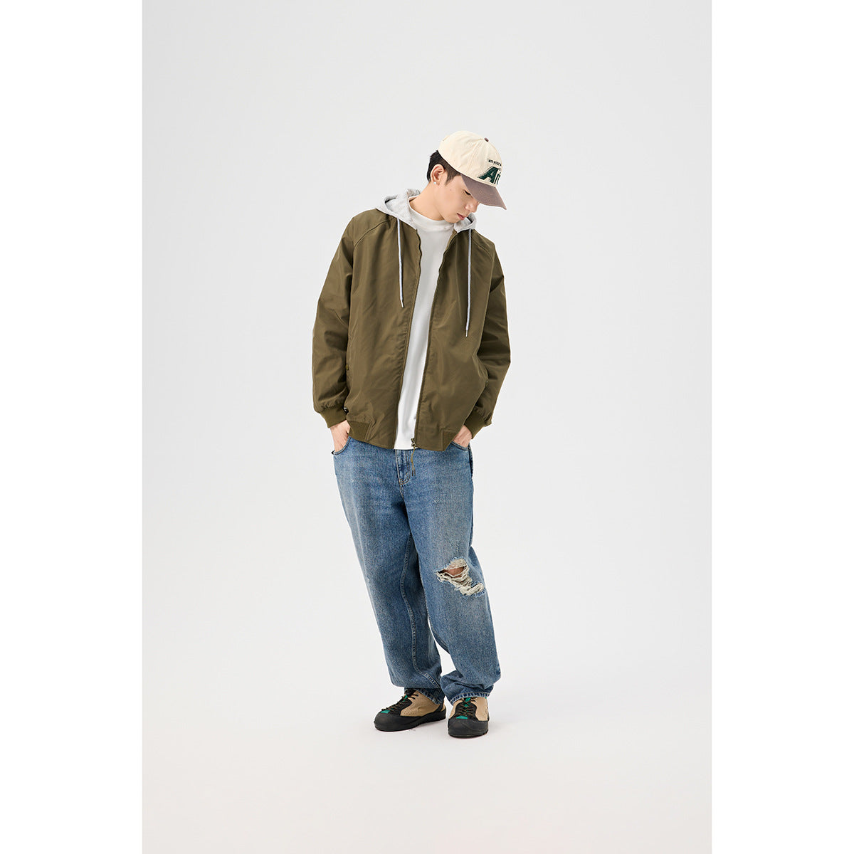 Model wearing an olive zip-front jacket with a gray hood, white shirt, loose blue jeans, and a beige cap.