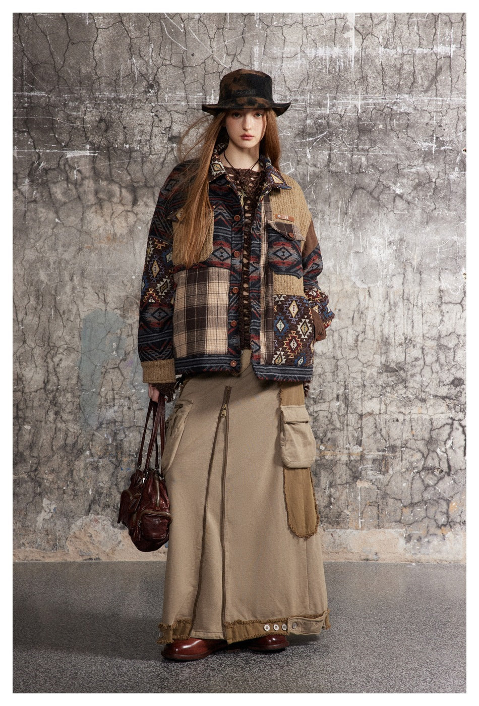 Model wearing the patchwork jacket open-styled with a hat, long khaki cargo skirt and brown leather bag against a gray wall