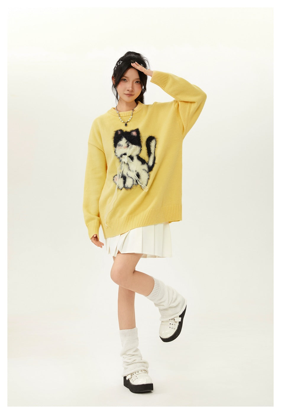Model poses in a yellow oversized sweater with a fuzzy cat graphic and a white pleated skirt.